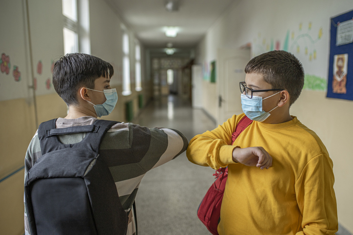 Zwei Schulkinder begrüßen sich in der Schule mit Masken und Elbogen.